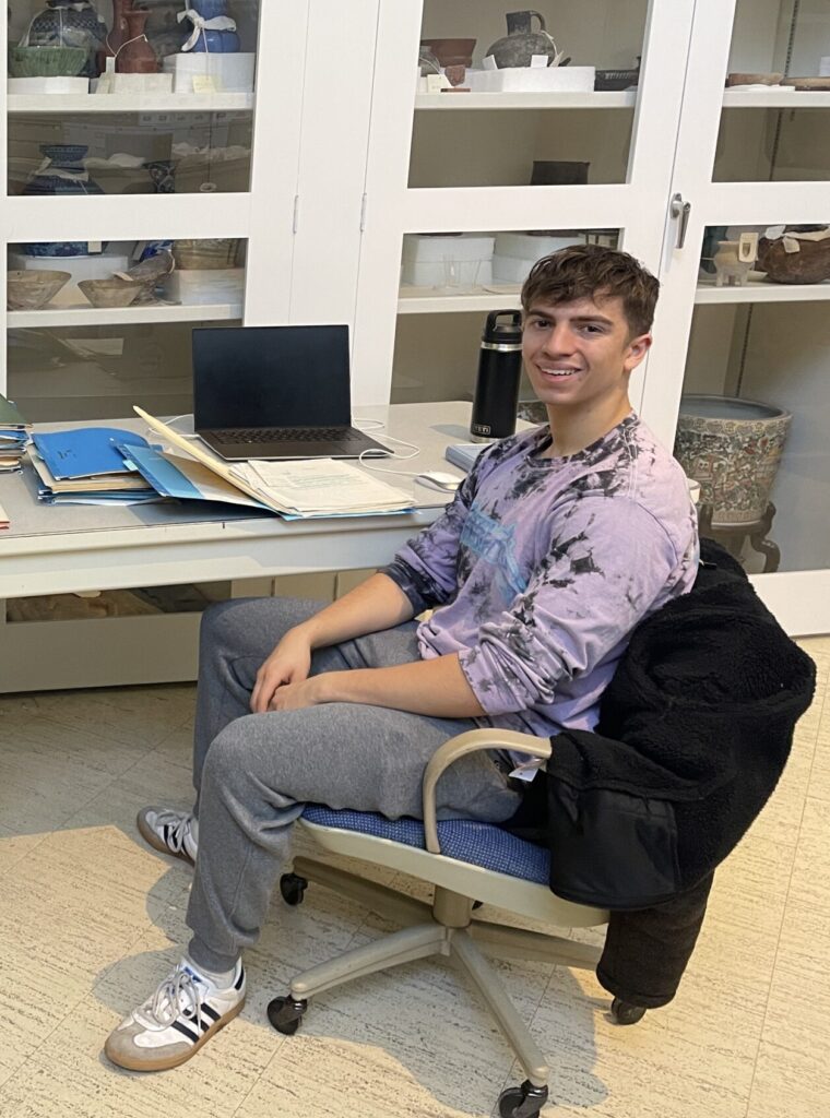 Two student interns sit on either side of files of archival papers in a museum storage area.