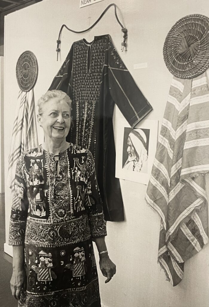 A black and white photograph shwoing woman wearing highly ornamented garb standing in front costume originating from the Middle East.
