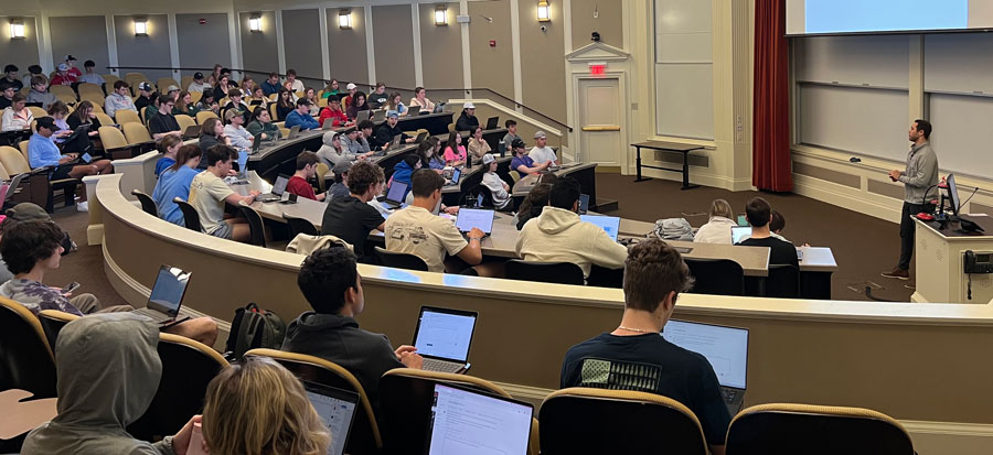 Students take notes on their laptops as they listen to a speaker addressing a large audience in an auditorium