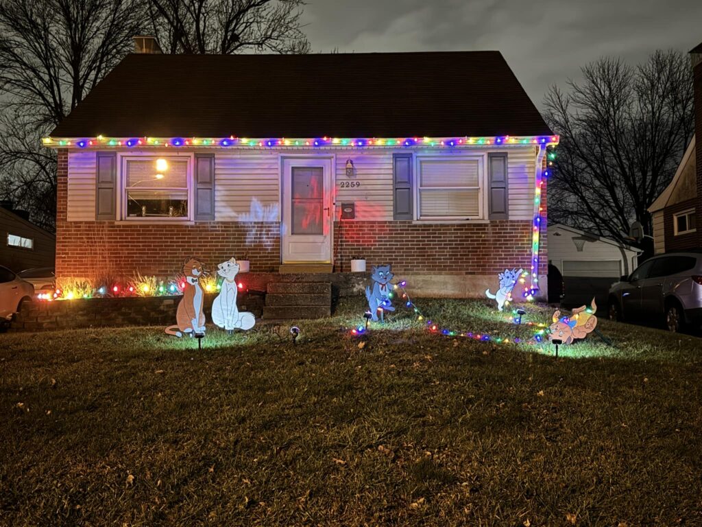 A brick house with holiday decorations. There are colorful light strings around the roof of the house and cutouts of the "Aristocats" in the yard - the kittens are displayed in a way that it makes them look like they are playing with the light strings.