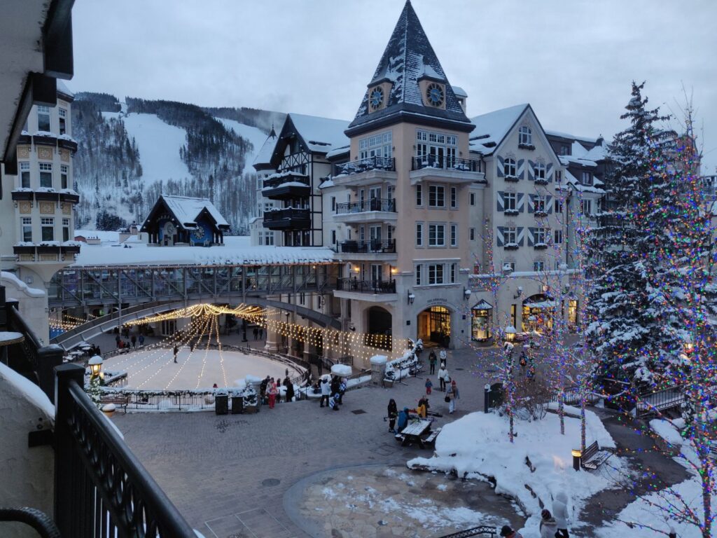 Vail, Colorado in the Winter. Tall, cream-colored buildings come to a point with holiday lights and snow-covered trees surrounding them. In the distance, you can see the snowy mountainside.