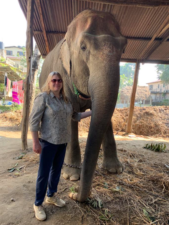 Michelle Szydlowski stands respectfully next to a large Asian elephant in a small enclosure in Nepal. The elephant has a chain around its front foot, and housing is visible in the background.