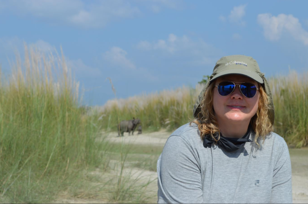 A smiling woman, Michelle Szydlowski, wears a hat and sunglasses in a tall grassland. In the background, under a bright blue sky, a mother rhinoceros and her baby are visible but softly out of focus.