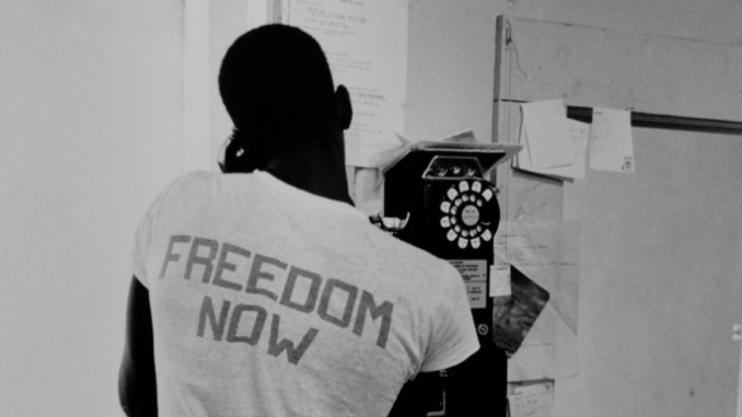 Photo of Freedom Now, "Summer of '64," Meridian, Mississippi. A Freedom Summer volunteer is on the phone with his back to the camera. His shirt says "FREEDOM NOW."