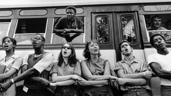 Photo of We Shall Overcome. Six protesters stand side-by-side, holding hands, and singing together.