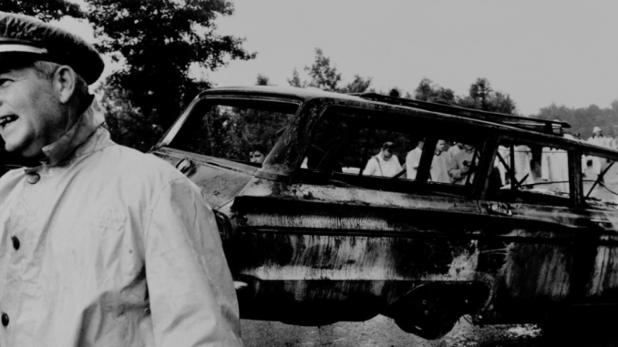 Photo of Civil Rights Worker's Station Wagon, Philadelphia, Mississippi. A policeman in the left corner is talking to someone off camera. In the background the burnt remains of a van hang.