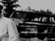 Photo of Civil Rights Worker's Station Wagon, Philadelphia, Mississippi. A policeman in the left corner is talking to someone off camera. In the background the burnt remains of a van hang.