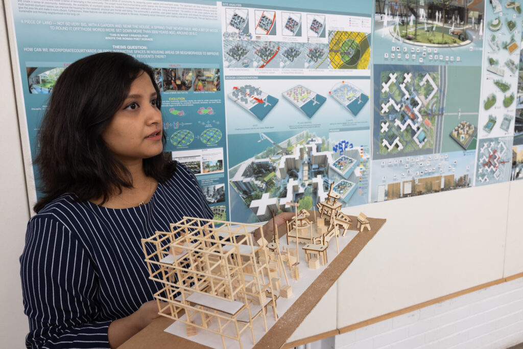 A student holds a wooden architectural model in front of a display board filled with design plans and conceptual renderings.