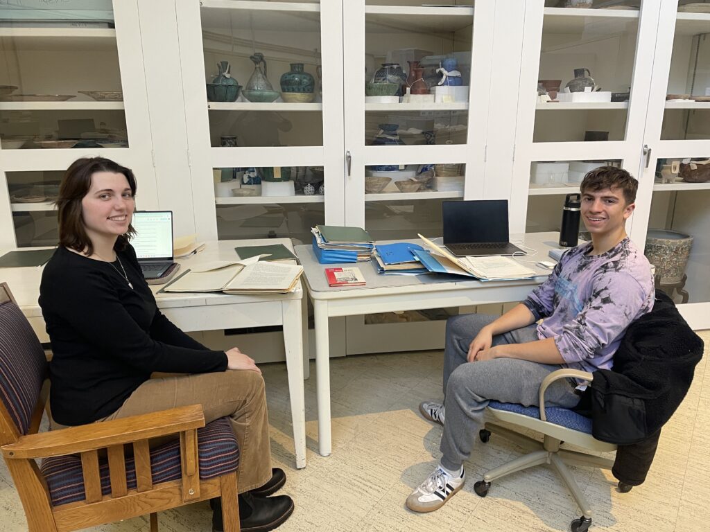 Two student interns sit on either side of files of archival papers in a museum storage area.