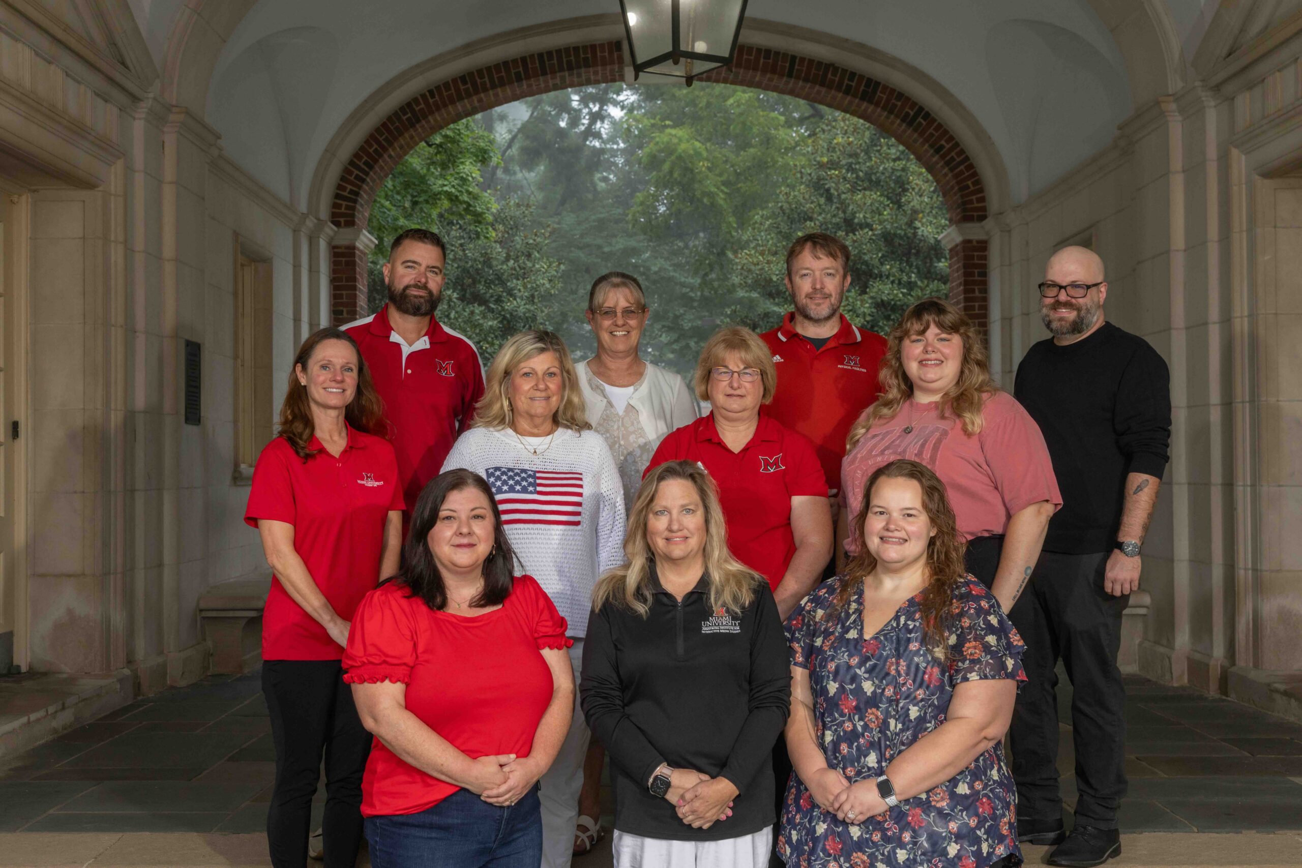 A group of ten individuals poses together under an arched stone walkway, with some wearing red shirts featuring a logo and one person holding an American flag.
