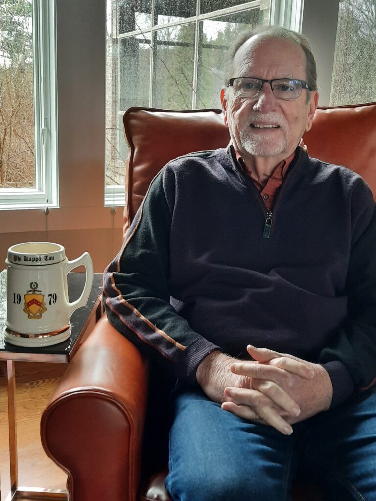 Photo of Bill Ash sitting in an armchair. To his right is a Phi Kappa Tau mug displaying the year 1979.