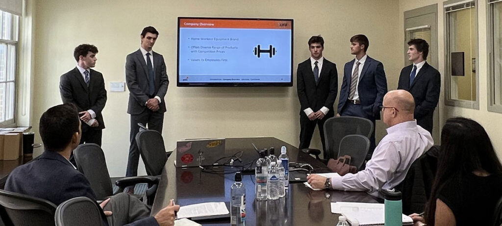 A team of five students presenting in front of a small panel seated at a conference table