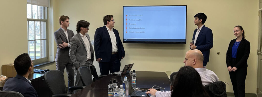 A team of five students gaze at their presentation on-screen others seated at a conference listen.