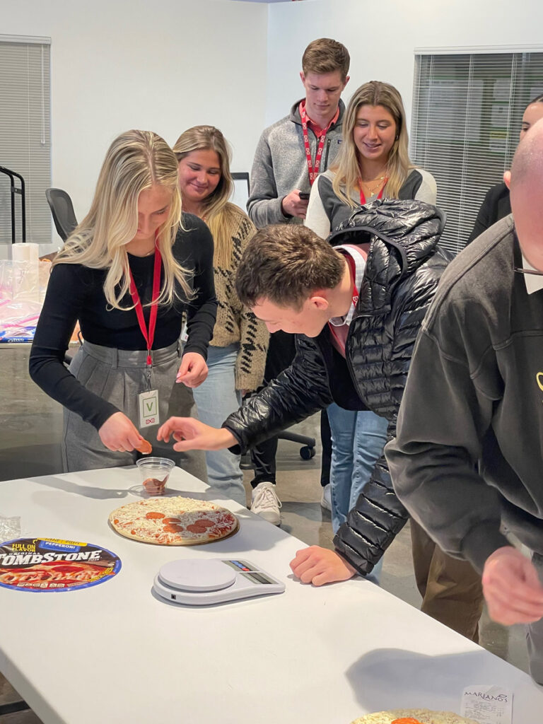 Students look on as as one of their peers examine a pizza on a table.