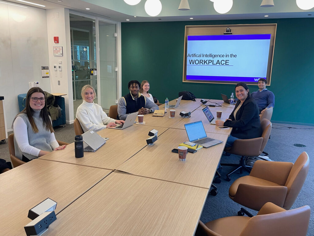 A team of students seated at a conference table with a slide presentation in the background