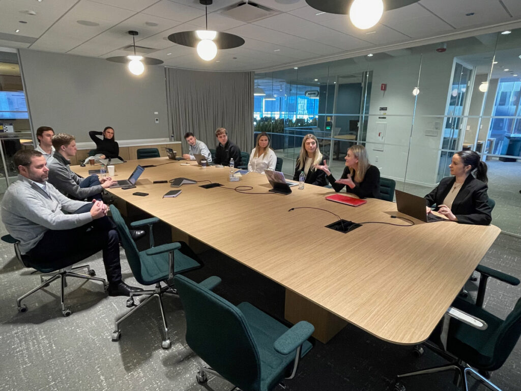 A long conference table with professionally dressed participants seated with their laptops having a discussion.