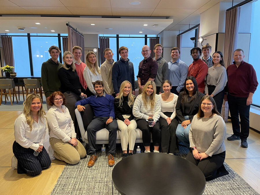 A group of college students pose with their professor inside an open office setting.