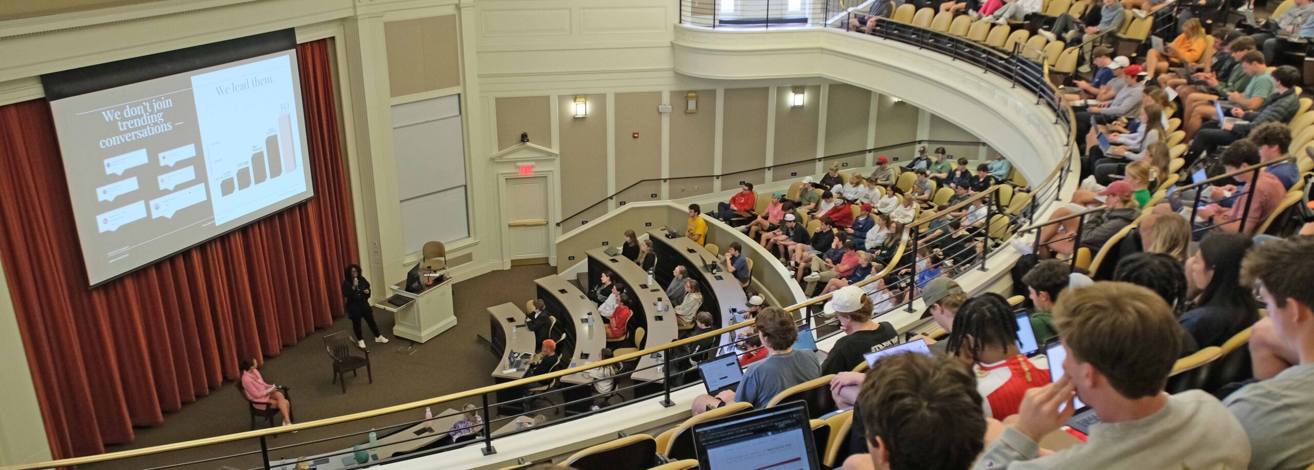 A wide-angle photo of a packed auditorium with two speakers on stage