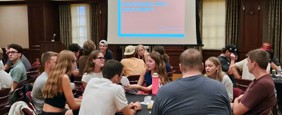 Photo of students seated at round tables with a slide projected in the background displaying the words "Professional Skills"