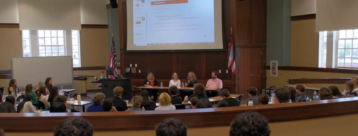 Photo of four panel members seated at a narrow table in front of a large auditorium audience