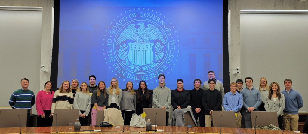 A group of students pose with Professor Eaton behind a large conference table, with the seal of the Board of Governors of the Federal Reserve System displayed on a screen behind the group.