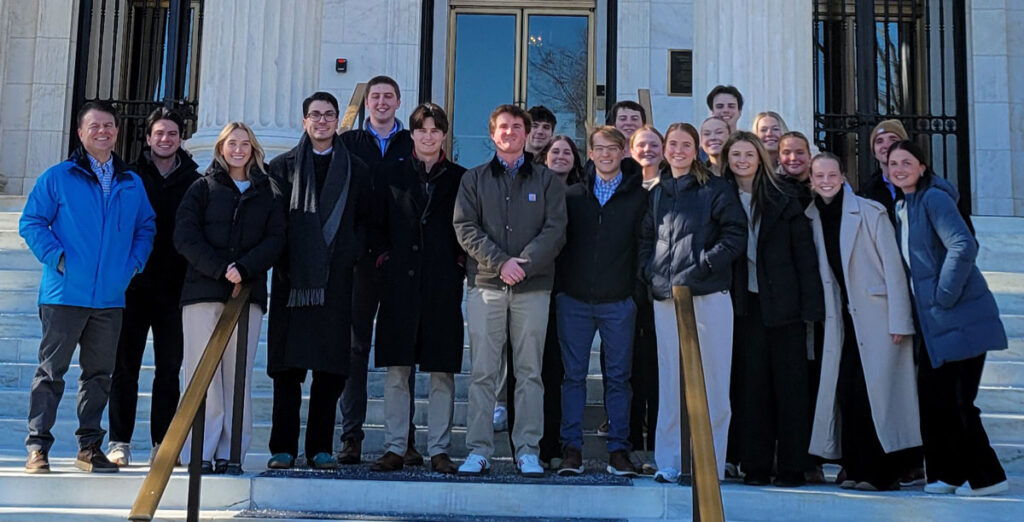 A group of Miami accountancy students with Professor Tim Eaton pose in front of a white brick building