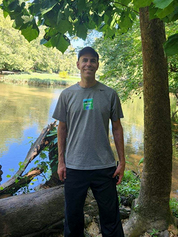 A photo of Greg during a hike outdoors, with water and shady trees in the background.