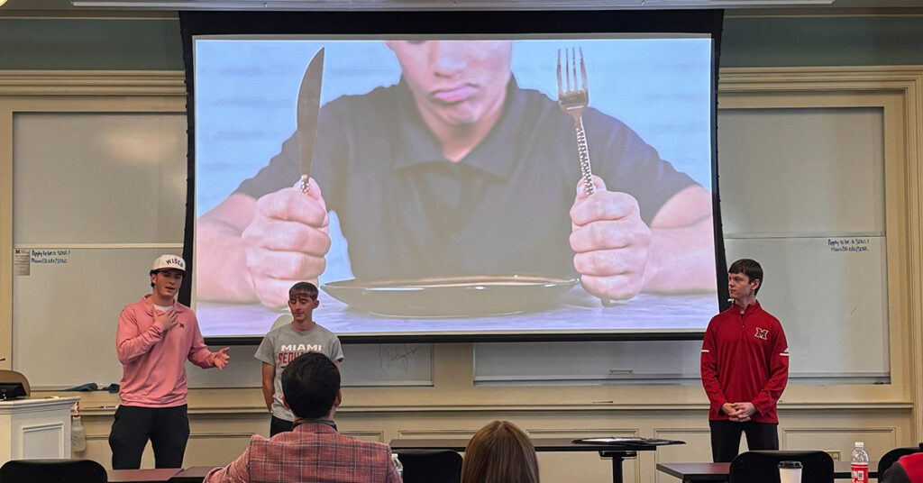 The winning team with a presentation slide in the background showing a close-up of a disgruntled man with an empty plate, holding a knife and fork.