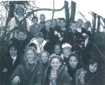 A black and white vintage-style photograph featuring a large group of approximately twenty smiling children and one adult woman huddled together on a wooded slope. They are all looking upward toward the camera lens against a background of bare trees. The photograph is currently oriented at a diagonal angle.