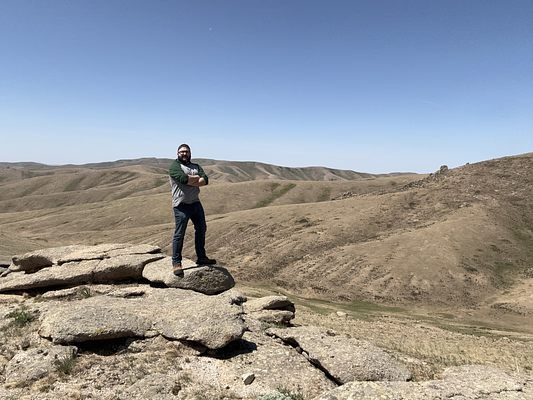 Dave Johnston standing atop a large rock formation in Mongolia, looking out over a vast, grassy landscape under a bright blue, cloudless sky.