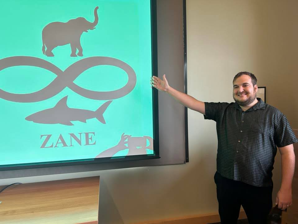 Grayson stands by a presentation screen displaying the ZANE logo.