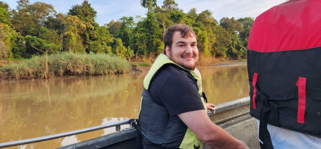 Grayson Ponti sits on a small boat on a river, wearing a life vest, with a backdrop of lush, green tropical forest in Borneo.