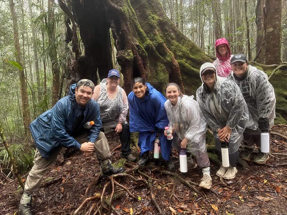 A smiling group of Project Dragonfly students standing outdoors in a forest, and wearing rain gear, during an Earth Expeditions graduate field course in Australia. 