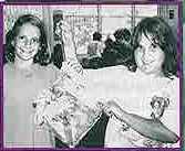 A black and white photograph framed with a magenta border. Two smiling young girls are shown in a classroom setting, holding up a clear plastic bag that contains a large leaf, demonstrating a plant transpiration experiment.