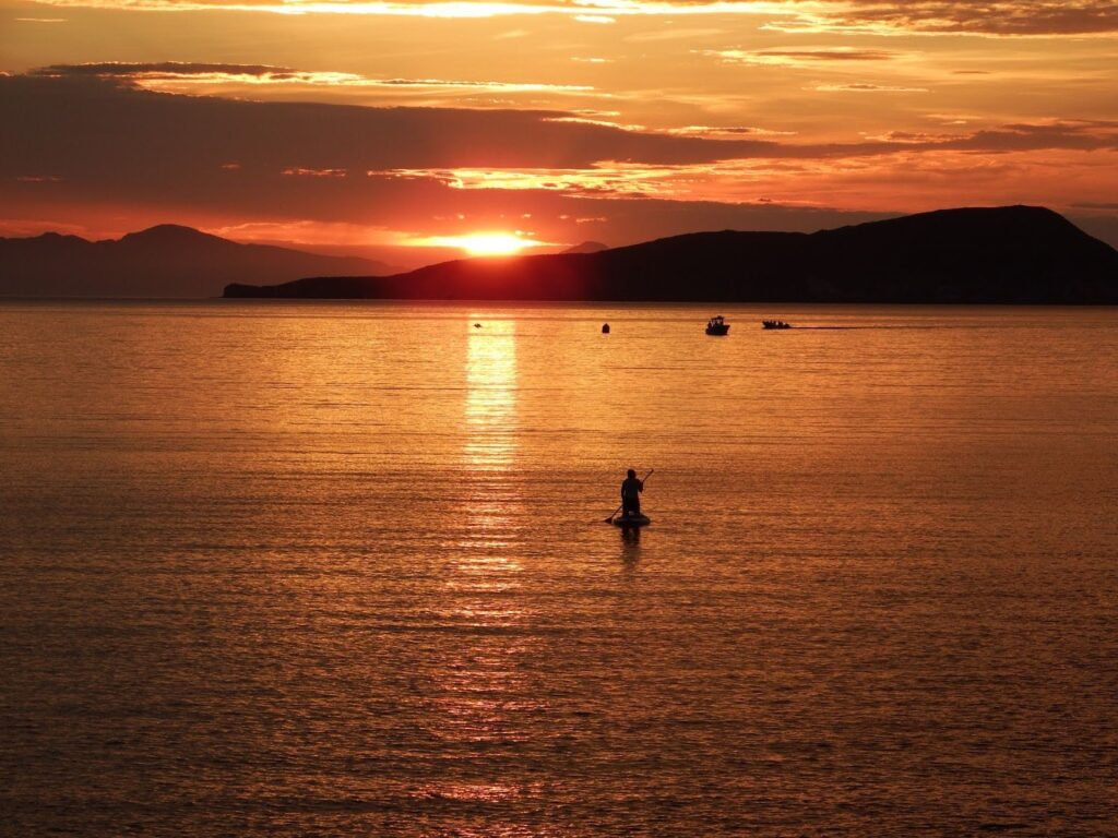 A wide landscape photograph capturing a vibrant sunset over the calm waters of the Gulf of California. Silhouetted islands rise in the distance against a sky filled with orange, deep pink, and blue hues.