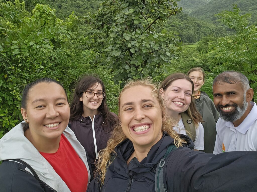 A close-up "usie" of Mikayla Deiotte smiling in the center, surrounded by five other Earth Expeditions participants, all smiling with a backdrop of dense tropical vegetation.