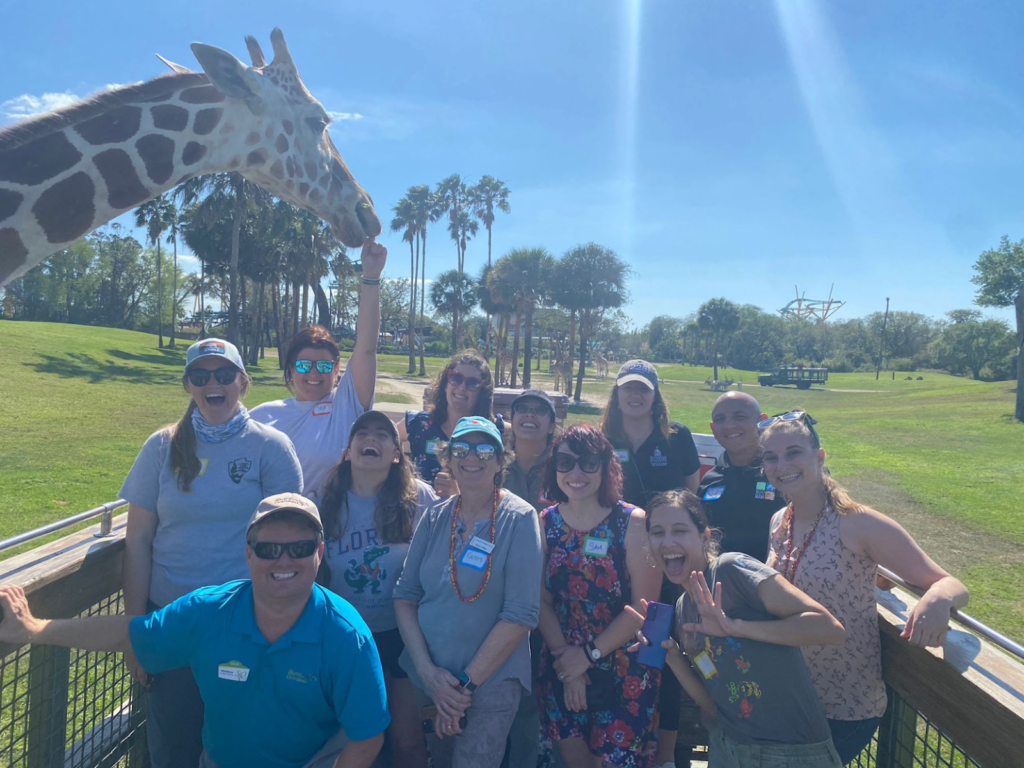 A group of smiling NNOCCI members posing together at Busch Gardens Tampa, with a giraffe visible in the background on the left.