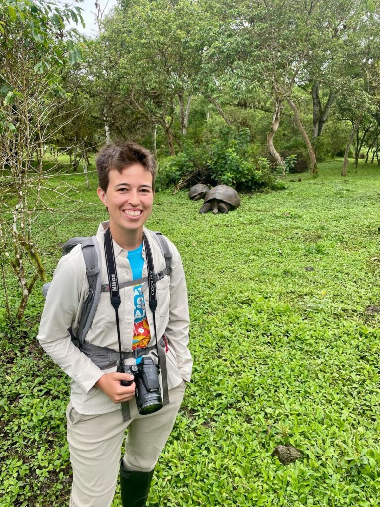 Cosette De Ferrari with a camera and backpack stands in a green field near giant tortoises.