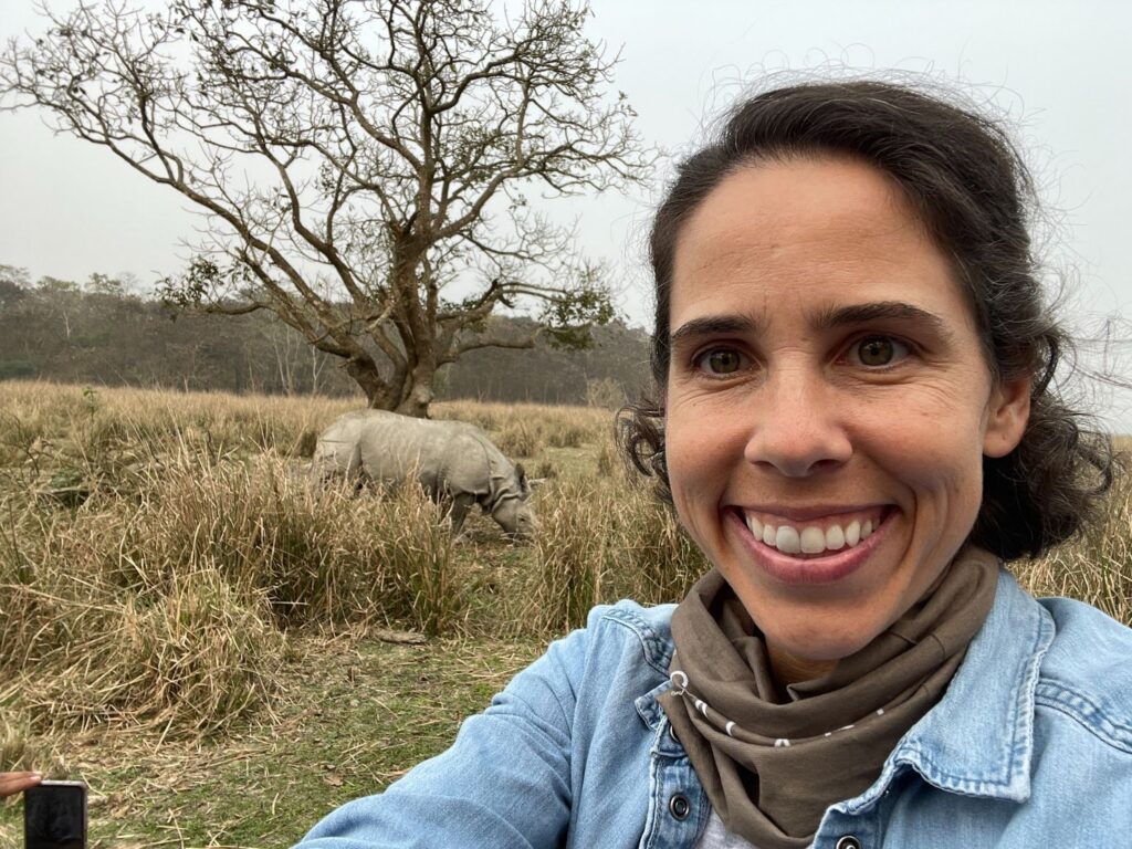 Martha Parker smiling in the foreground with a large, armored Greater One-Horned rhino grazing in the tall grass behind her in India.