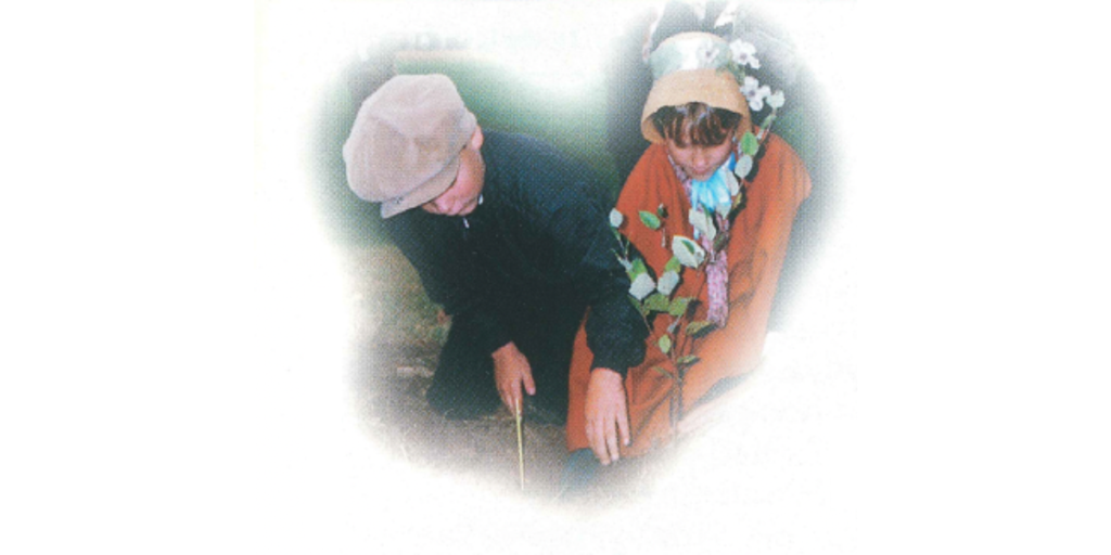 A vintage photograph showing two young students dressed in 1800s-style historical costumes, including a flat cap and a shawl. They are kneeling on the ground outdoors, carefully planting a small tree sapling as part of a school history project.