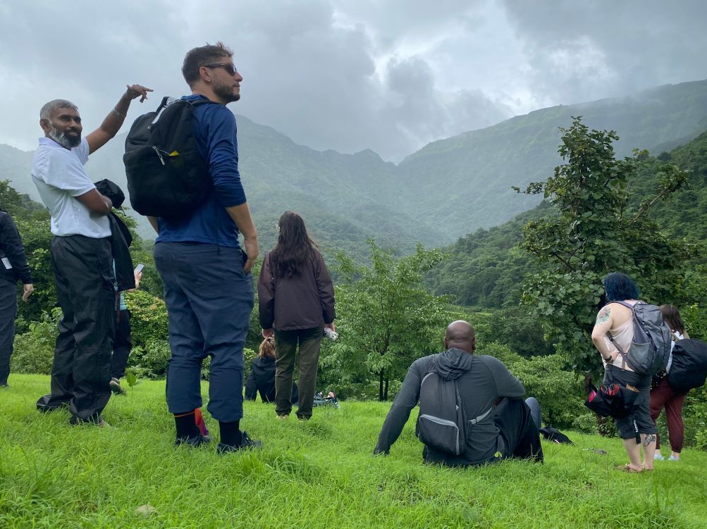 A group of students seen from behind, some sitting on the grass and some standing, looking out over a vast landscape of misty mountain ridges and deep green valleys in India.