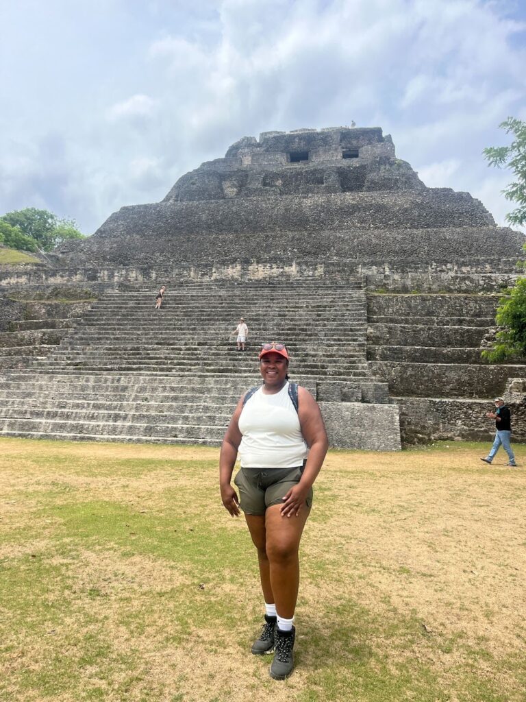 A photo of Lyndeisha Curry, a woman with a bright smile, standing on grassy ground in front a pyramid at Xunantunich, Belize, under a blue sky with some clouds.