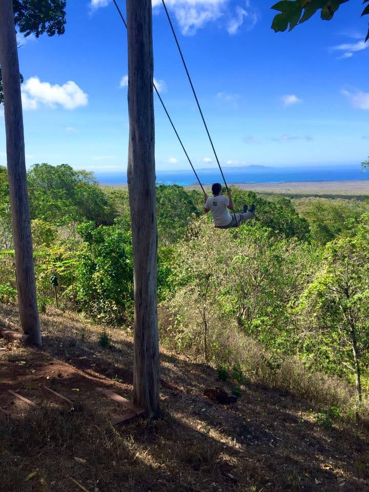 Martha Parker on a high wooden swing, soaring out over a vast, green valley and sunny landscape in the Galápagos Islands.