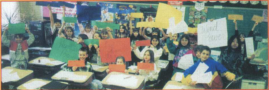 Students sit at a school desk using string and small wooden dowels to construct a hanging mobile featuring cutouts of flying objects.