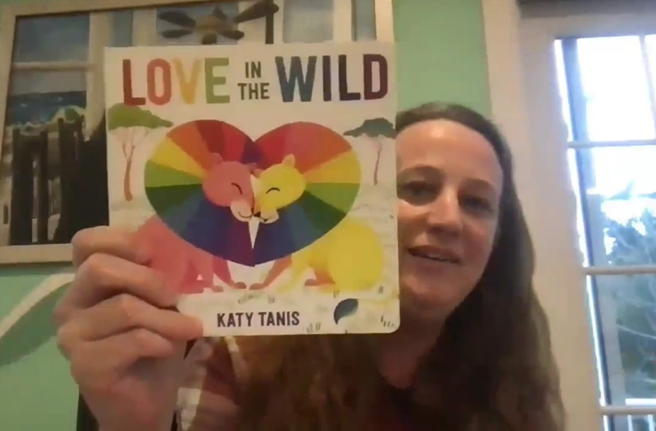 An image of Katy Tanis smiling and holding her colorful board book titled "Love in the Wild," which features two lions with their manes forming a heart.