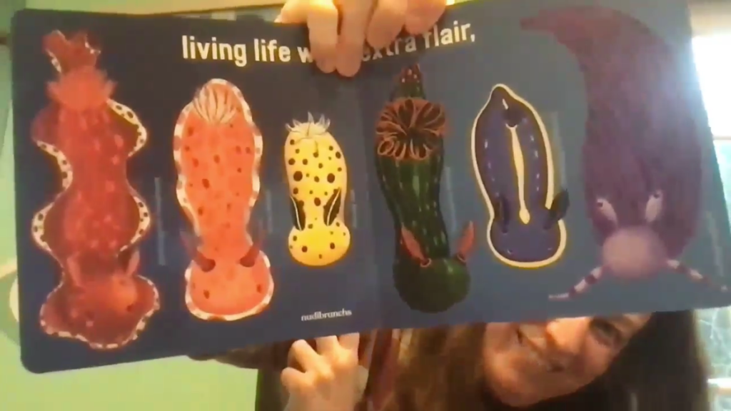 Katy Tanis smiles from behind her open book, "Love Under the Sea." The page spread shows several brightly colored sea slugs (nudibranchs) with the text "living life with extra flair."