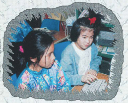 Two young girls, one in a colorful tie-dye shirt and one in a blue cardigan, working together at a computer.