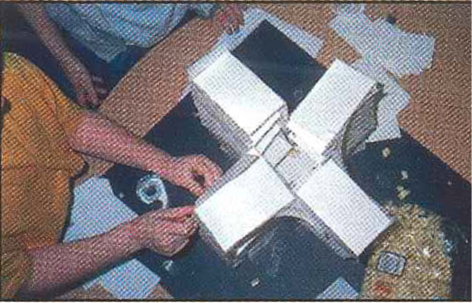 An overhead view of a white index card colony built in the shape of a cross with four distinct wings.