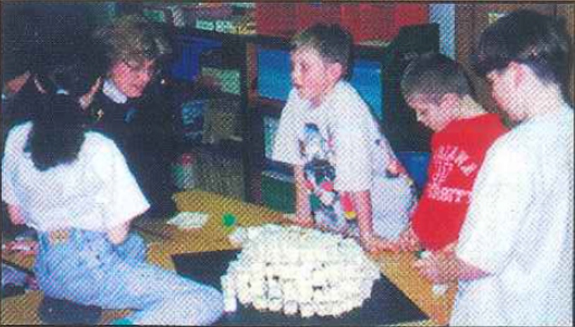 Four fourth-grade students collaborating to build a complex structure out of white index cards on a classroom table.
