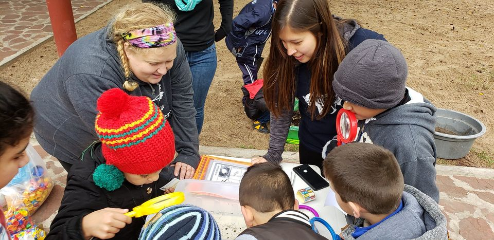 Two educators and a group of young students gathered outdoors around a learning table. The children are highly engaged, using bright red and yellow magnifying glasses to examine natural objects while the teachers smile and guide the hands-on science activity.